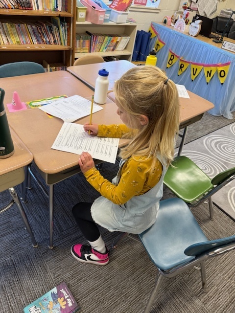 A young girl sits at a classroom desk, focused on completing a worksheet with a pencil.