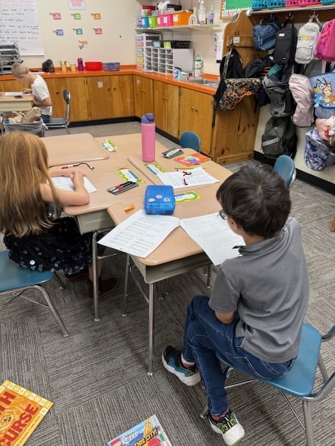 Two young students sit at a classroom table, working on worksheets with school supplies spread out in front of them.