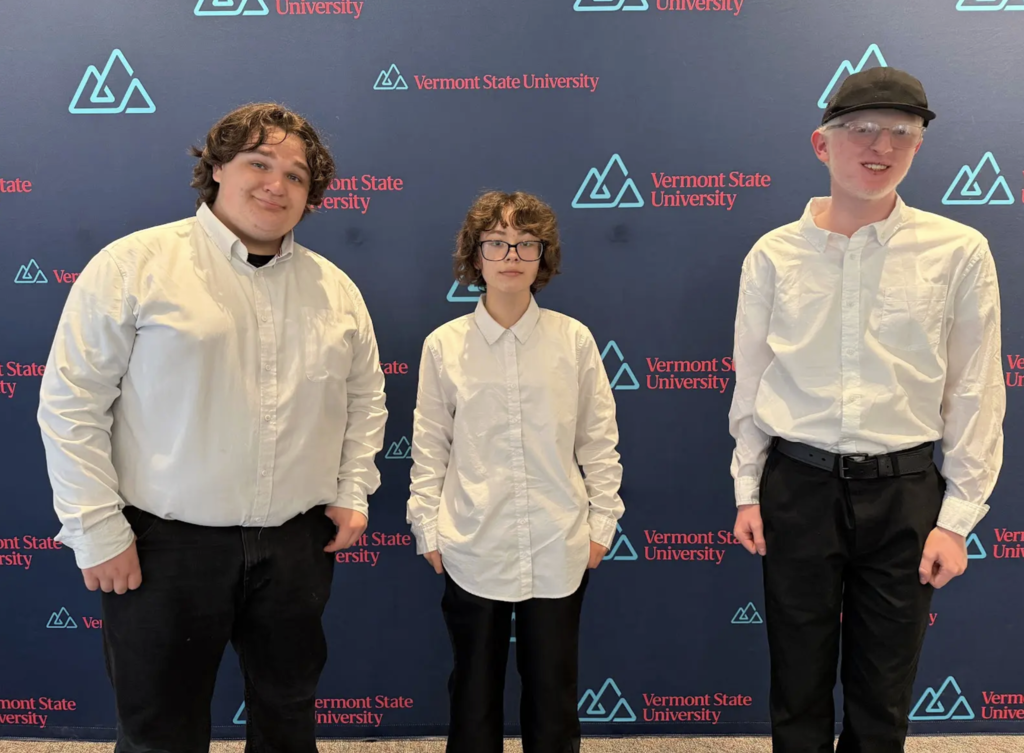 Three students dressed in white button-down shirts and black pants stand in front of a Vermont State University step-and-repeat backdrop. The student on the left smiles with hands in pockets, the student in the middle stands with hands at sides, and the student on the right wears glasses and a black cap, smiling.