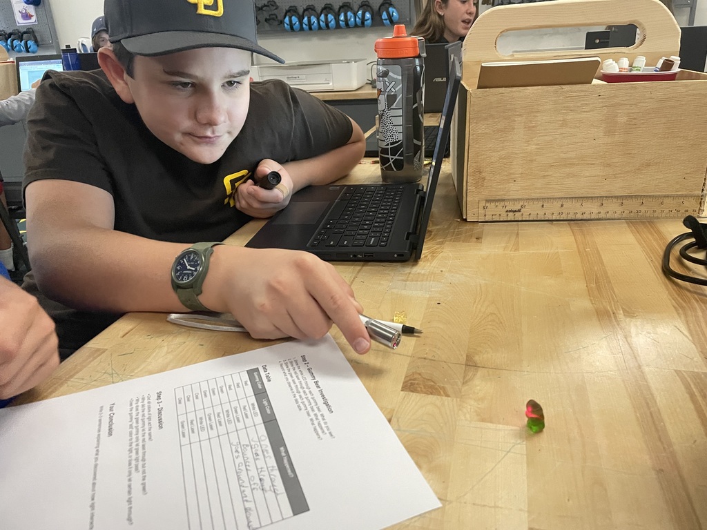 Student in a black shirt and cap points a laser light at a gummy bear on the table, with a worksheet nearby.