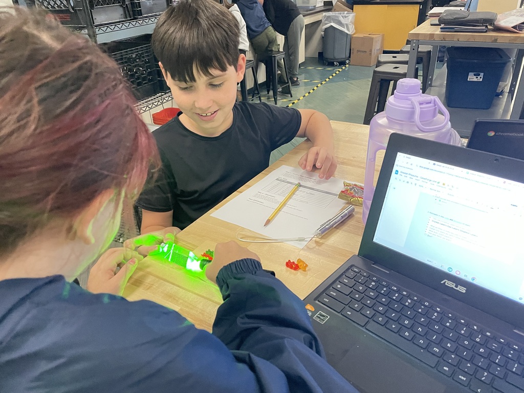 Two students at a table, one shining a green laser light on gummy bears while the other smiles and observes.