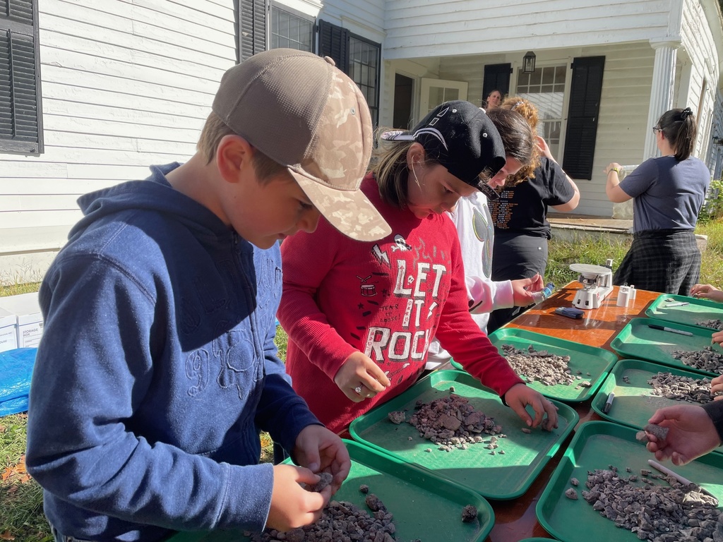 Students sort through dirt and rocks on green trays outside a historic building.