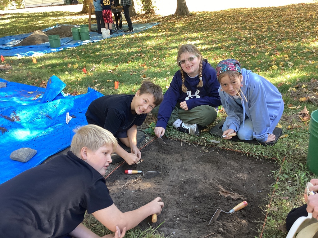 Four students kneel at a dig site, using small tools to excavate soil.
