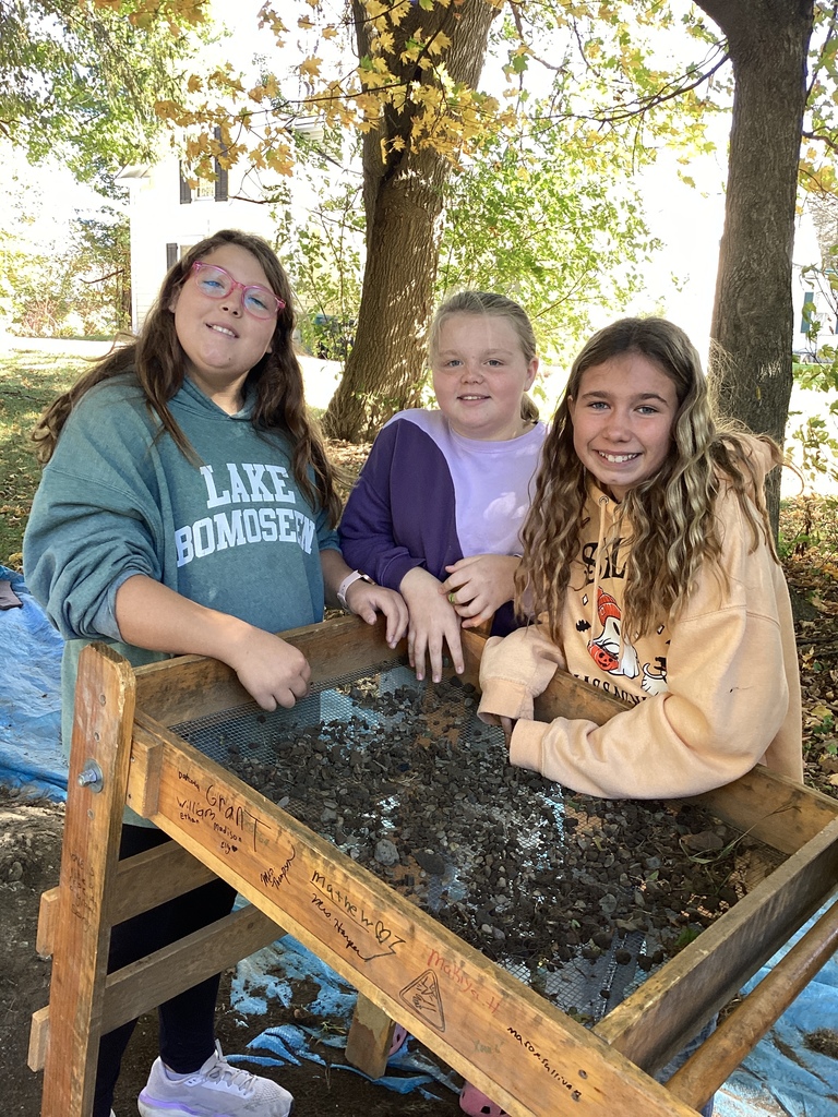 Three students smile while standing behind a sifting screen with dirt and artifacts.