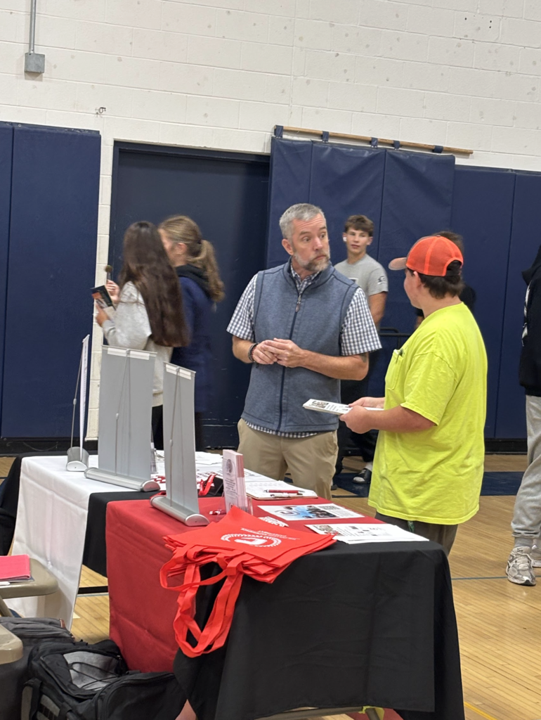 A man at a booth speaks with a student in a yellow shirt during a school career fair; tables display pamphlets and tote bags.