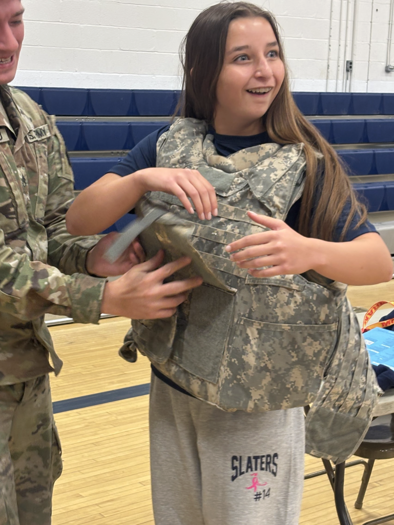 A student smiles while trying on a military camouflage vest with help from a service member.