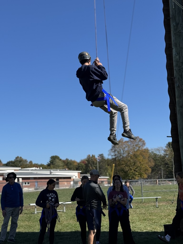 Student in helmet and harness is lowered on a rope, with others on the ground guiding.