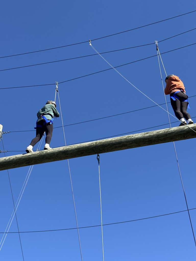 Two students in harnesses walk carefully across a high ropes log while clipped into safety lines.