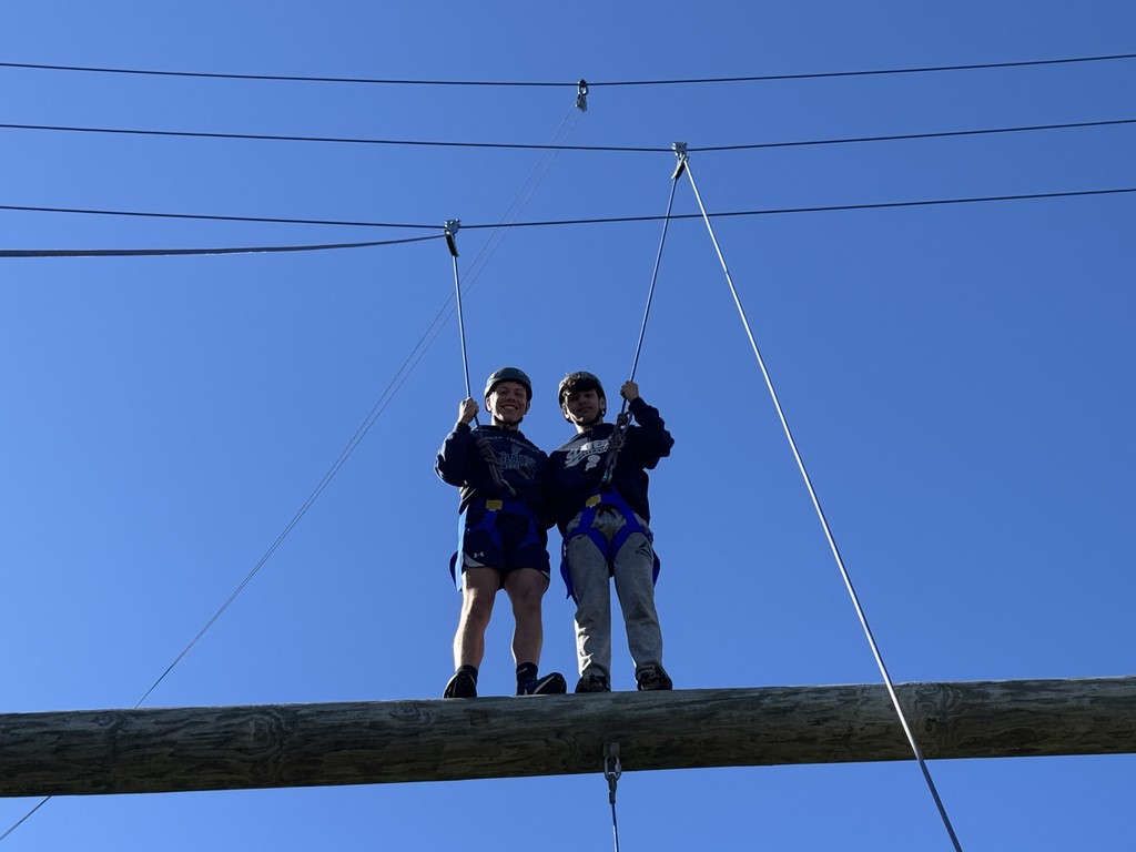 Two students in harnesses stand on a high ropes log element, smiling and holding safety lines against a blue sky.