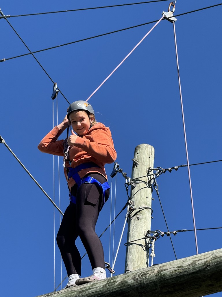 Smiling student in an orange sweatshirt gives a thumbs up while standing on a high ropes log.
