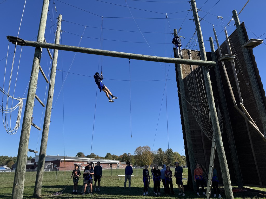 Student descends on a rope from a high ropes course as classmates and staff watch from below.