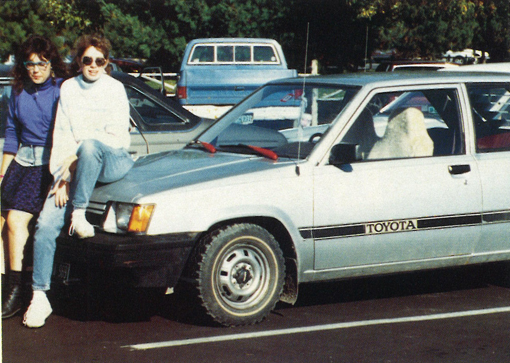 Two students sitting on the hood of a silver Toyota parked in a school lot, smiling at the camera.