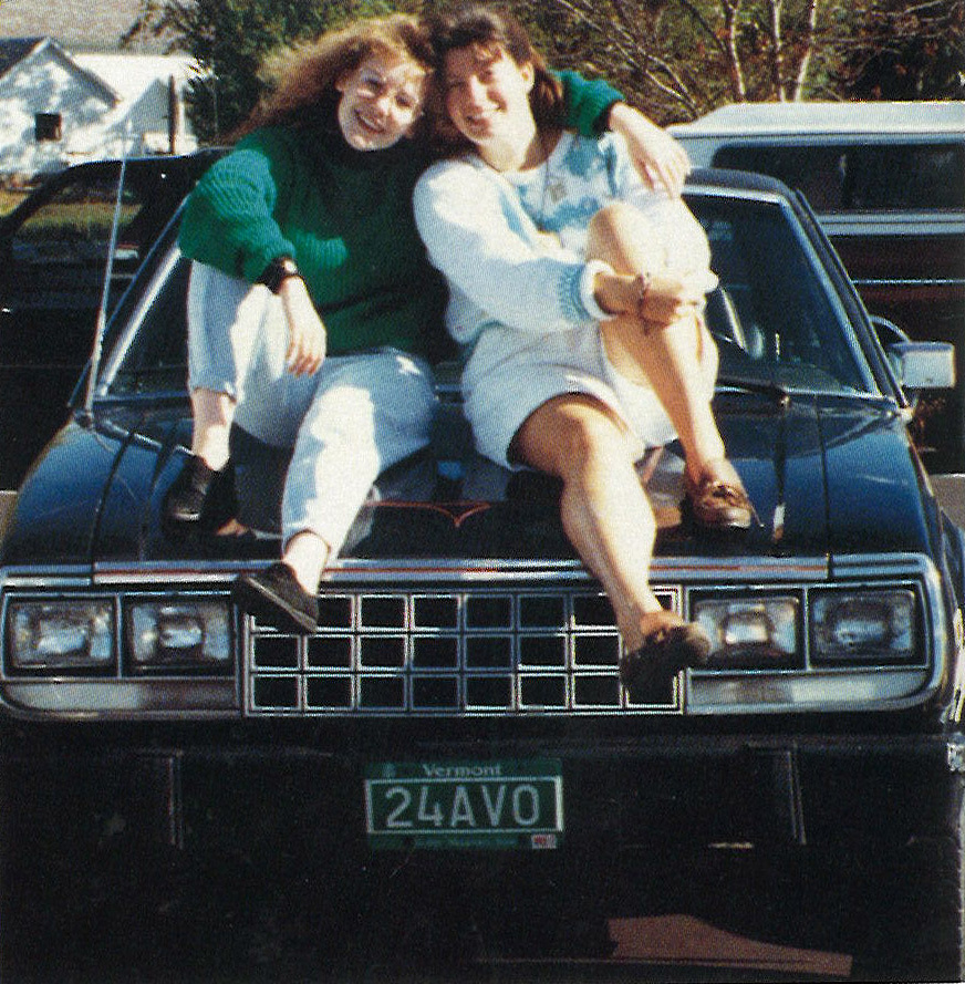 Two friends sitting on the hood of a dark car with a Vermont license plate, laughing and posing together.