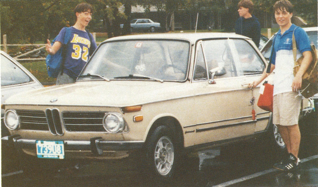 Two students standing beside a beige car in a school parking lot, one wearing a Lakers shirt, smiling and gesturing.