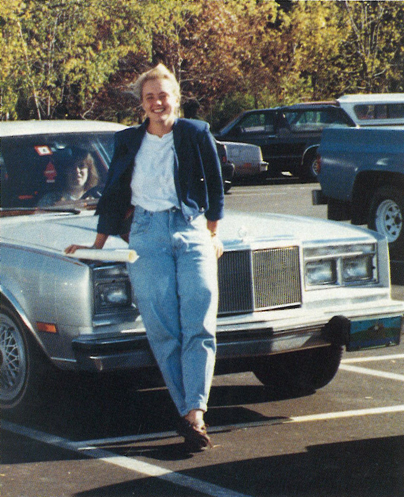 Student in a 1990s parking lot leaning against a silver car, smiling, wearing jeans and a blazer.