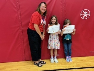 Teacher poses with two smiling students holding certificates, standing against a red gym mat backdrop.