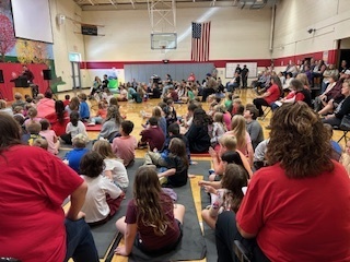 Students and staff gathered in a school gymnasium for an assembly, sitting on mats and chairs facing the front where presenters are speaking.