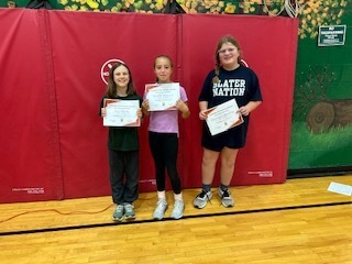 Three students hold certificates and smile for the camera in front of a red gym mat wall.