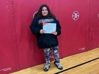 Student stands alone holding a certificate, smiling in front of a red gym mat wall.