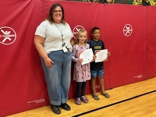 Teacher stands with two students holding certificates during a recognition event.