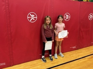 Two students stand side by side in front of a red gym wall, one holding a certificate.