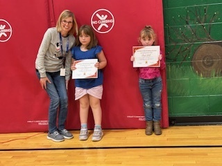 Teacher poses with two young students holding certificates during an assembly.