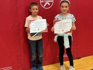 Two students stand side by side, each holding a certificate in front of a red gym mat wall.