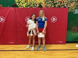 Two students pose together holding certificates, smiling in front of a red gym mat wall.