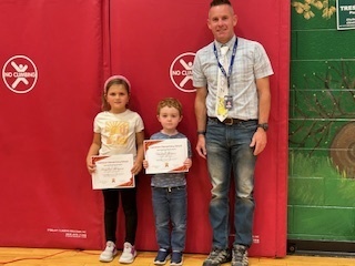 Two young students stand with a staff member, holding certificates in front of a red gym mat wall.