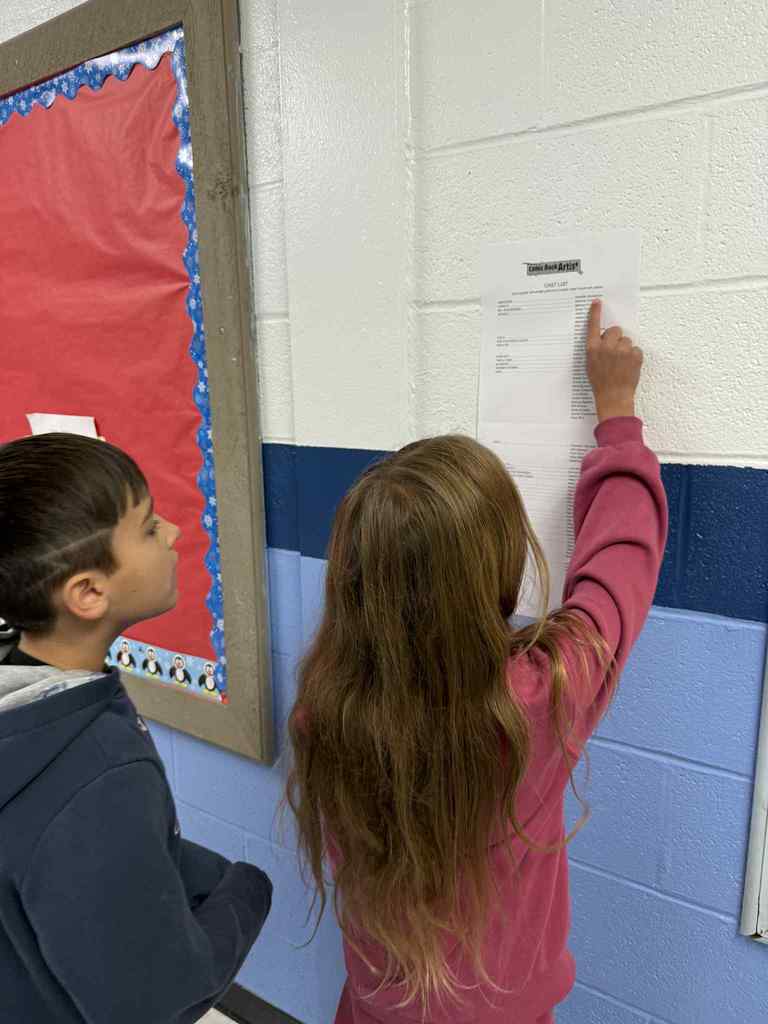 Two students stand in a hallway reading a paper on the wall, with one student pointing to a name on the list.