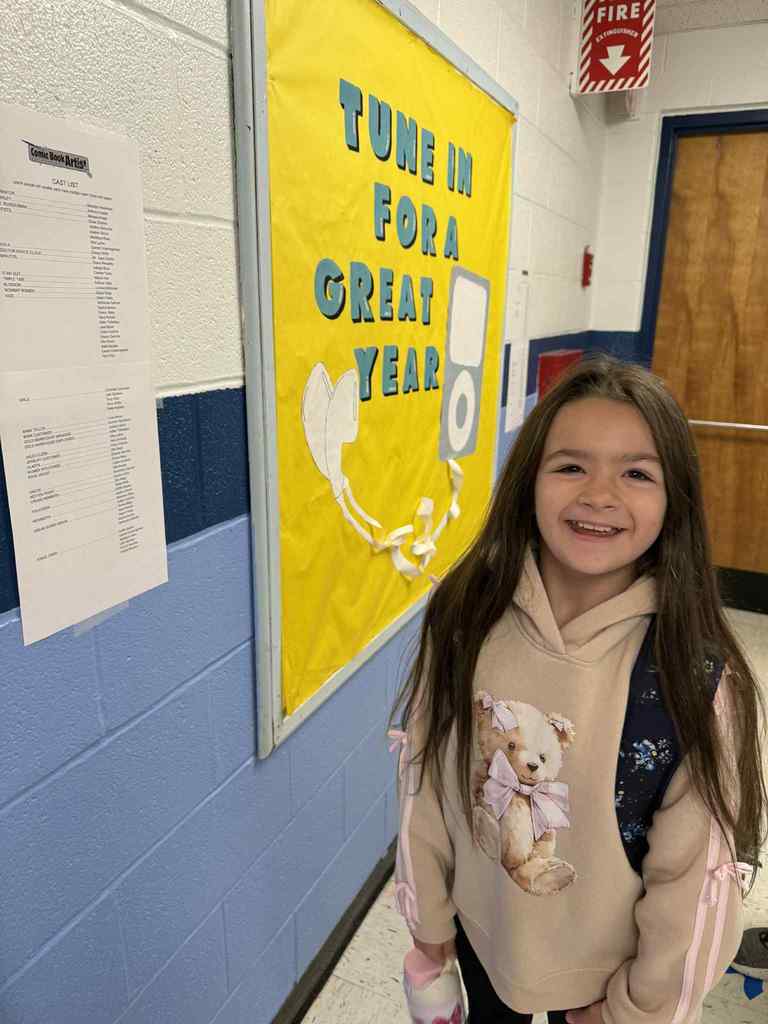 A smiling young girl with long brown hair stands in a school hallway. She is wearing a tan hoodie with a teddy bear design and a dark backpack. A yellow bulletin board behind her reads "TUNE IN FOR A GREAT YEAR" with an MP3 player graph