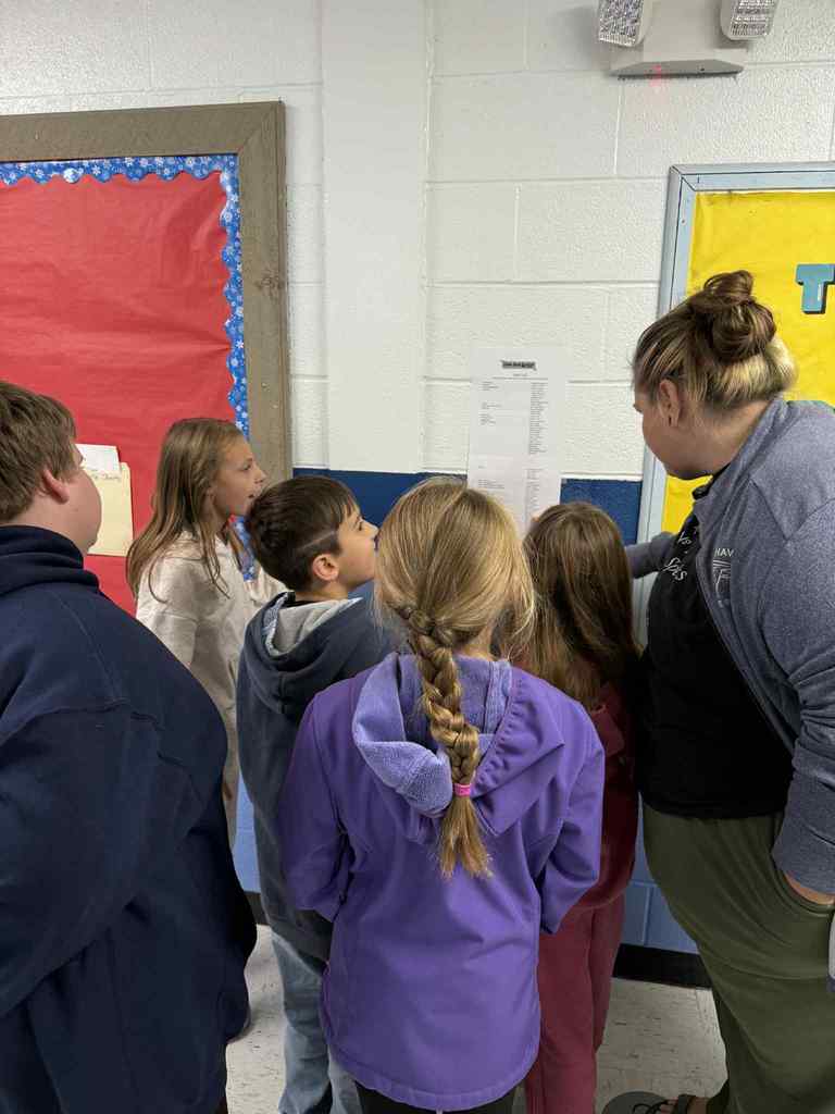 A teacher and several students stand in a hallway looking at a paper posted on the wall.