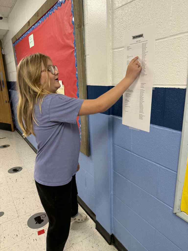 A young girl with blonde hair and glasses stands in a school hallway, wearing a light blue t-shirt and black pants, actively reading or pointing at a long, white "Cast List" titled "Comic Book Artists" posted on the blue wall.