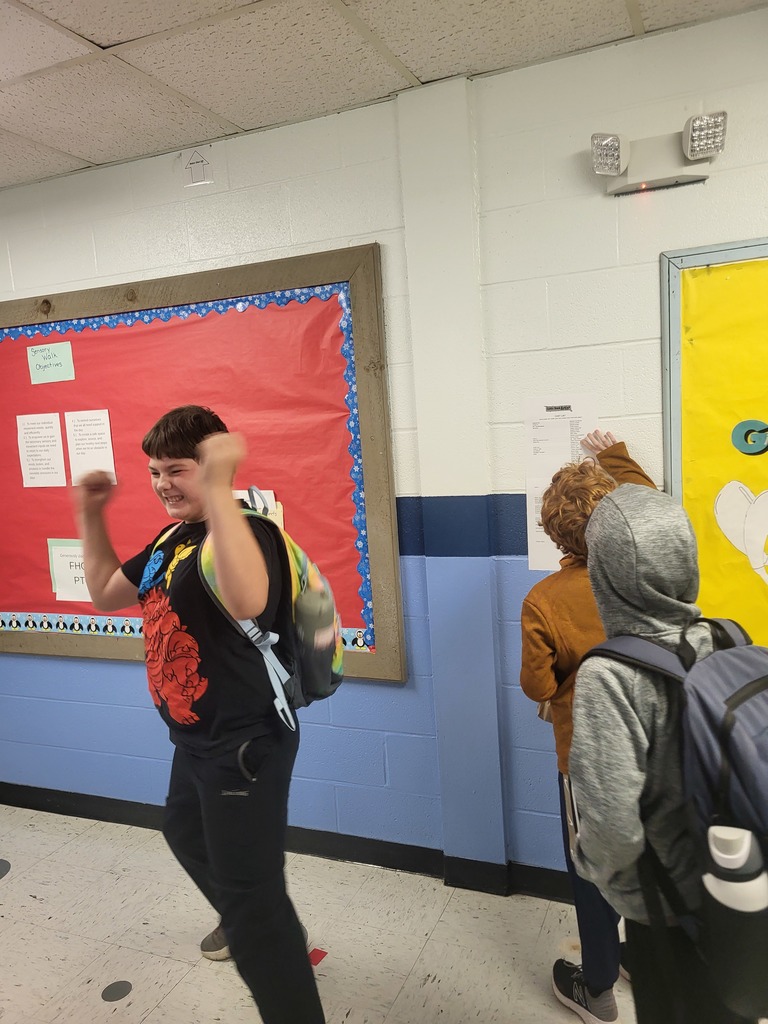 student cheering with hands in the air while other students are looking at the list of names on the paper on the wall.