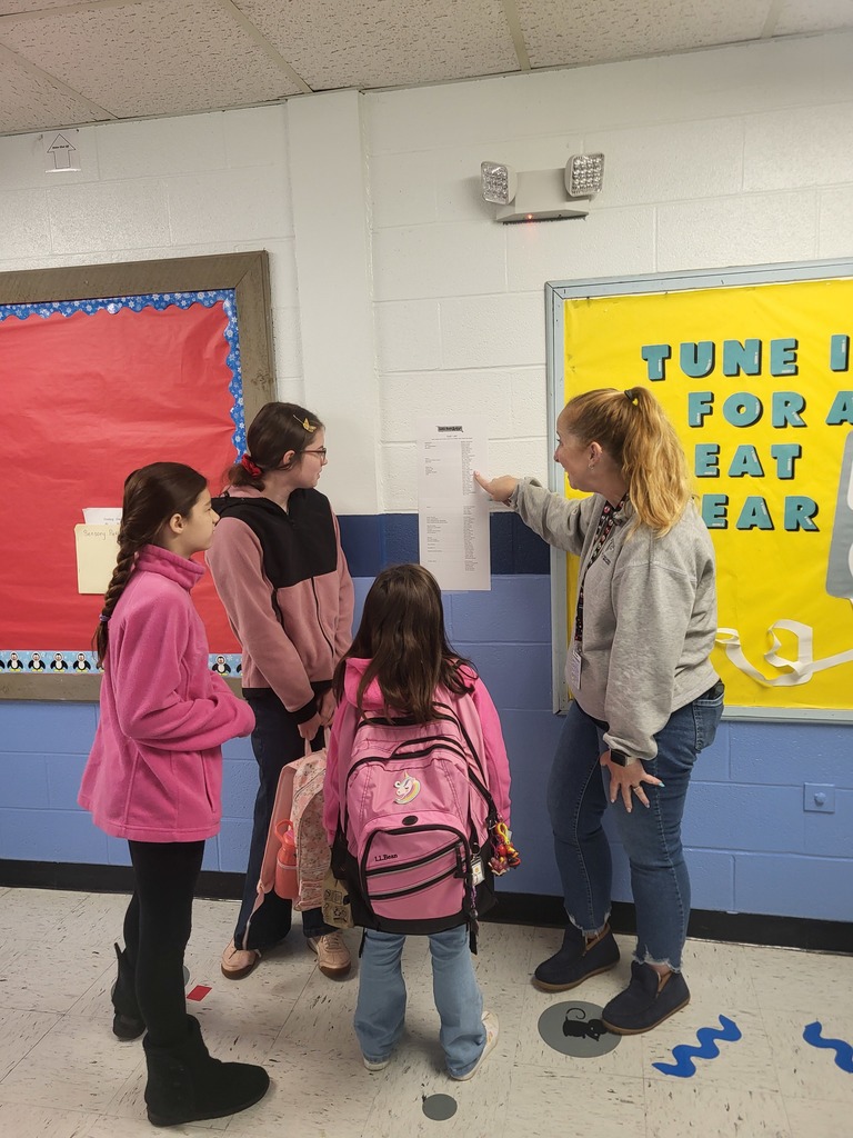 A teacher and three students stand in a hallway looking at a paper posted on the wall, with the teacher pointing to a spot on the list.