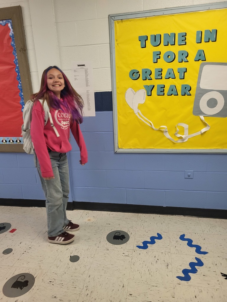 A smiling young girl with pink and purple dyed hair stands in a school hallway. She is wearing a pink hoodie with a "COBRA" logo, light-wash jeans, a white backpack, and dark sneakers. To her right is a large yellow bulletin board that says, "TUNE IN FOR A GREAT YEAR" next to a drawing of a classic MP3 player with headphones. The floor has circular animal print and blue wavy decals.