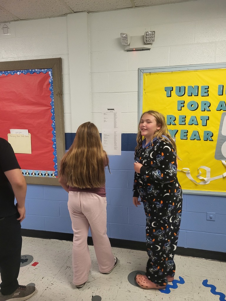 Two young girls are in a school hallway near a yellow bulletin board. One girl, with long brown hair, is facing away in pink flared pants. The other, with blonde hair, faces forward in black celestial-themed pajamas and pink slides. The bulletin board reads "TUNE IN FOR A GREAT YEAR."