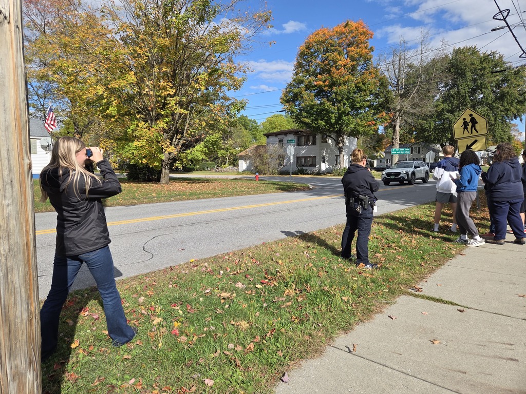 Students and an officer watch cars and run radar and lindar on a neighborhood street lined with colorful fall trees.