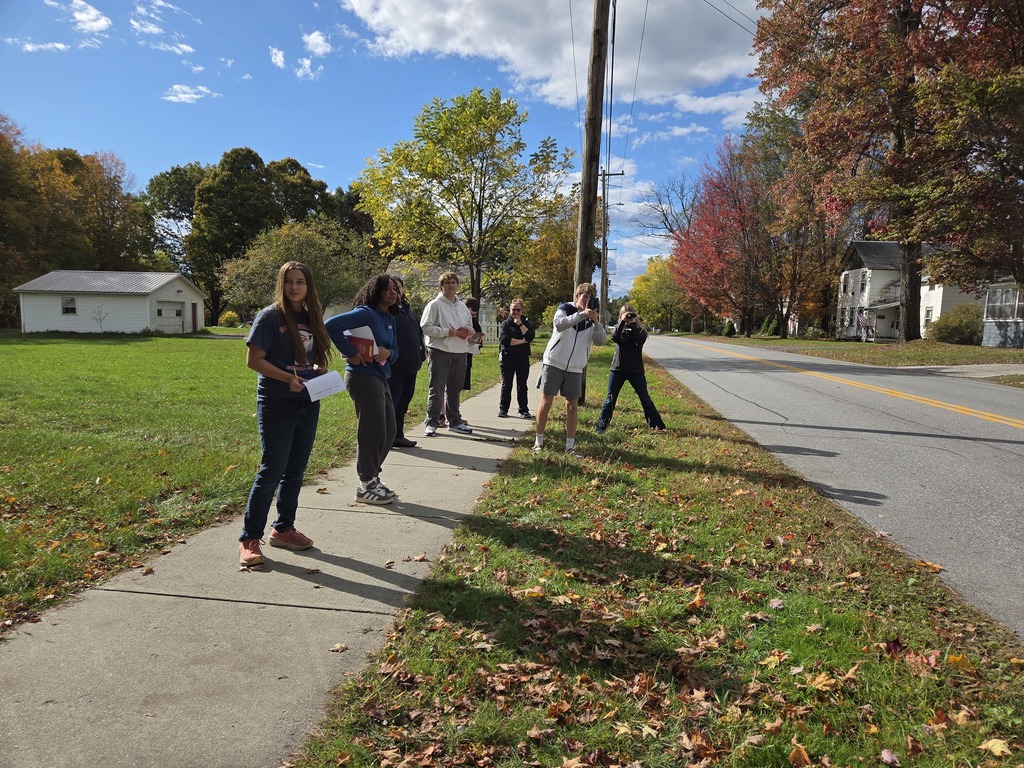 Students spread along a sidewalk with notebooks, watching traffic during a class activity under bright autumn trees.
