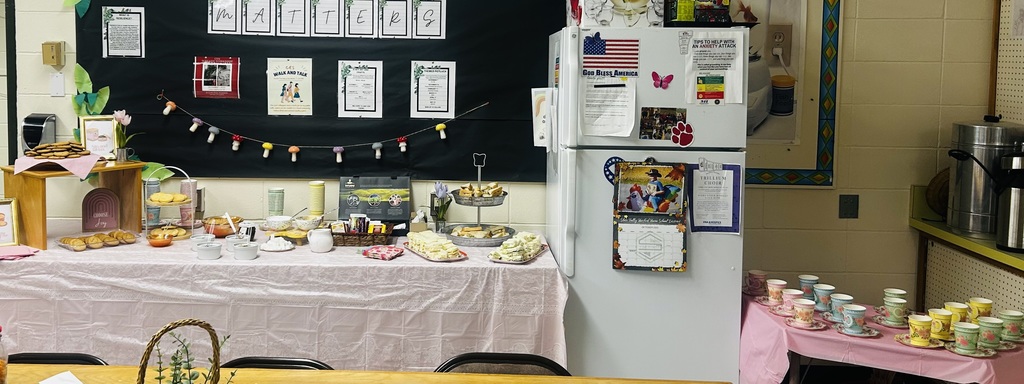 A table set up for a staff tea party with pink tablecloths, teacups, pastries, sandwiches, and framed decorations in a school break room.