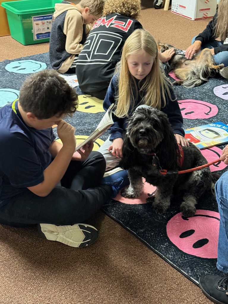 Students sit on a smiley-face rug reading books and petting two therapy dogs in a classroom.