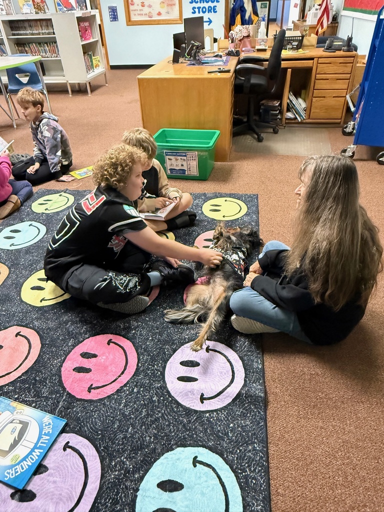 Students sit on a smiley-face rug reading books and petting a therapy dog in the library.