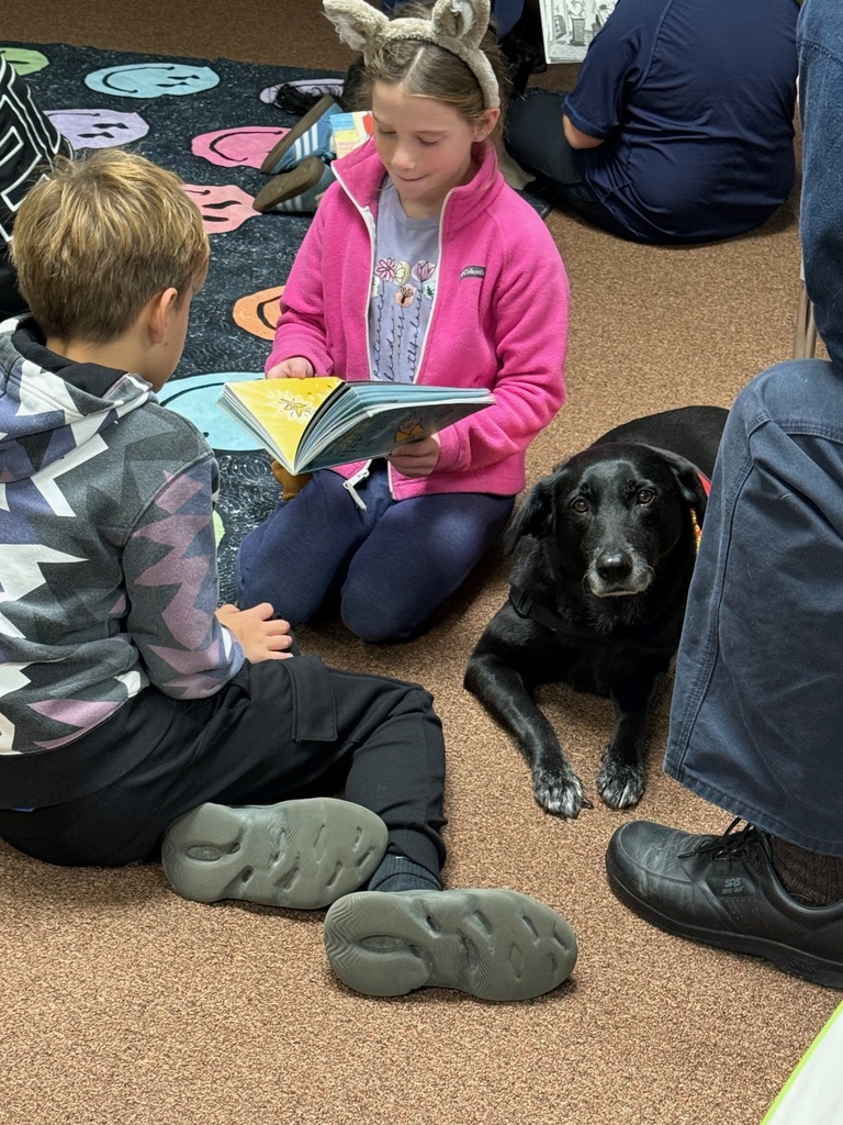 girl in a pink jacket holding a book and sitting next to a boy in a dark sweatshirt. A black dog is lying calmly on the floor next to them.
