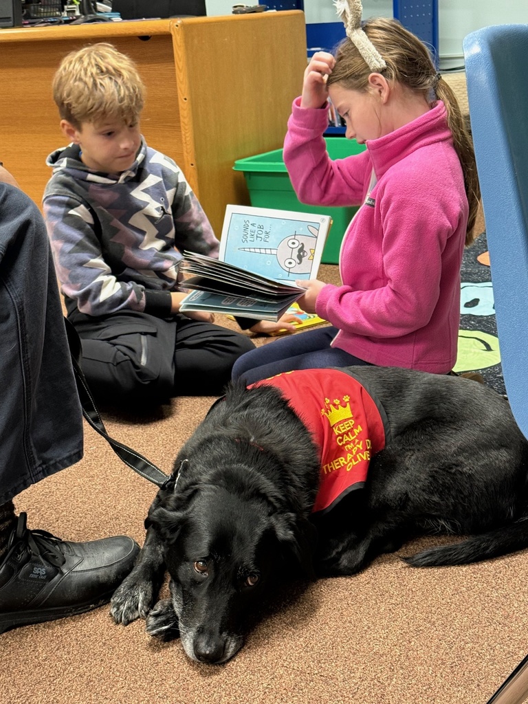 Two children sit on the floor reading books next to a black therapy dog wearing a red vest that says “Keep Calm and Read to Me.”