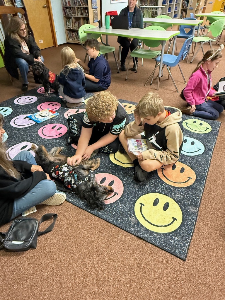 Students sit on a colorful smiley-face rug reading and petting therapy dogs during a reading session.