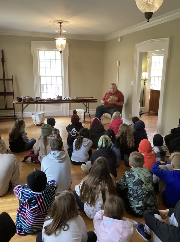 A large group of students sit on the floor listening to a guest speaker presenting cultural tools and items.