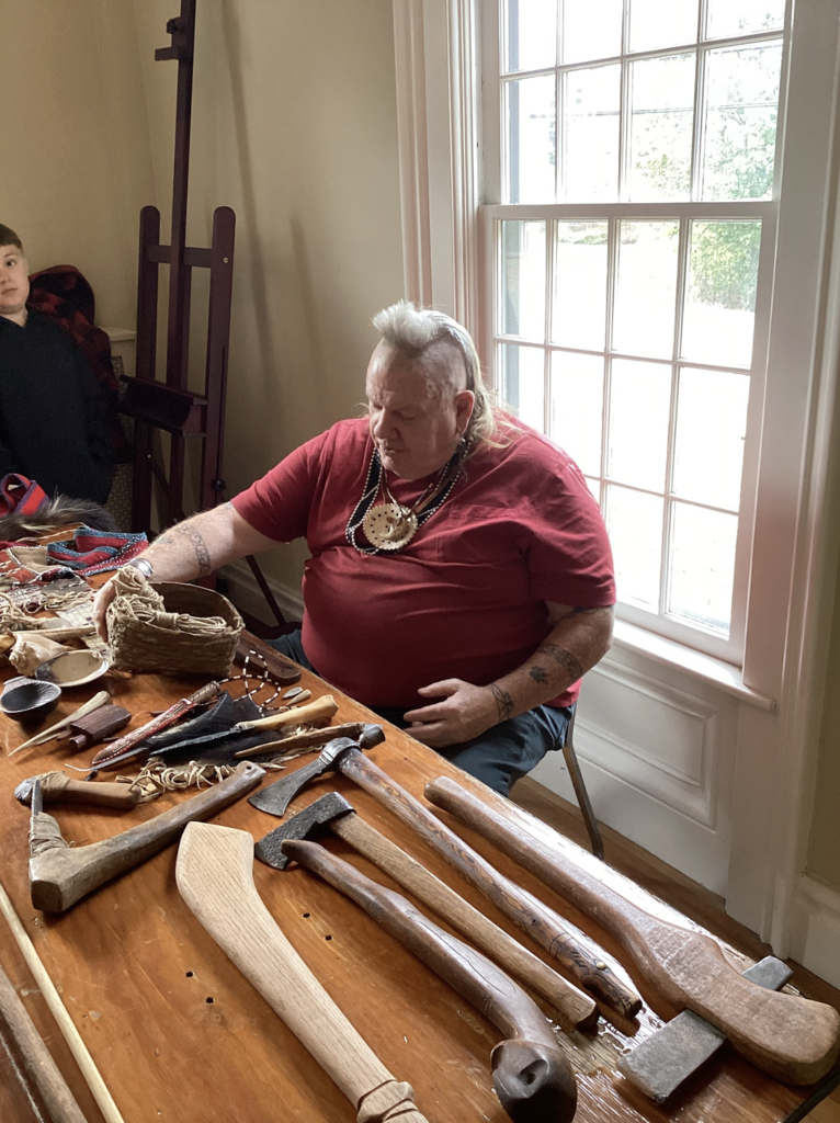 A presenter sits at a table displaying traditional hand tools and crafts, speaking to nearby students.