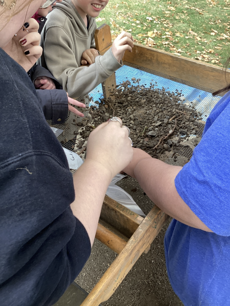 Close-up of students examining soil and small rocks on a mesh sifter during an excavation project.