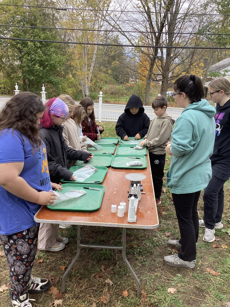 Students stand at an outdoor table using green trays and scales to analyze soil samples.