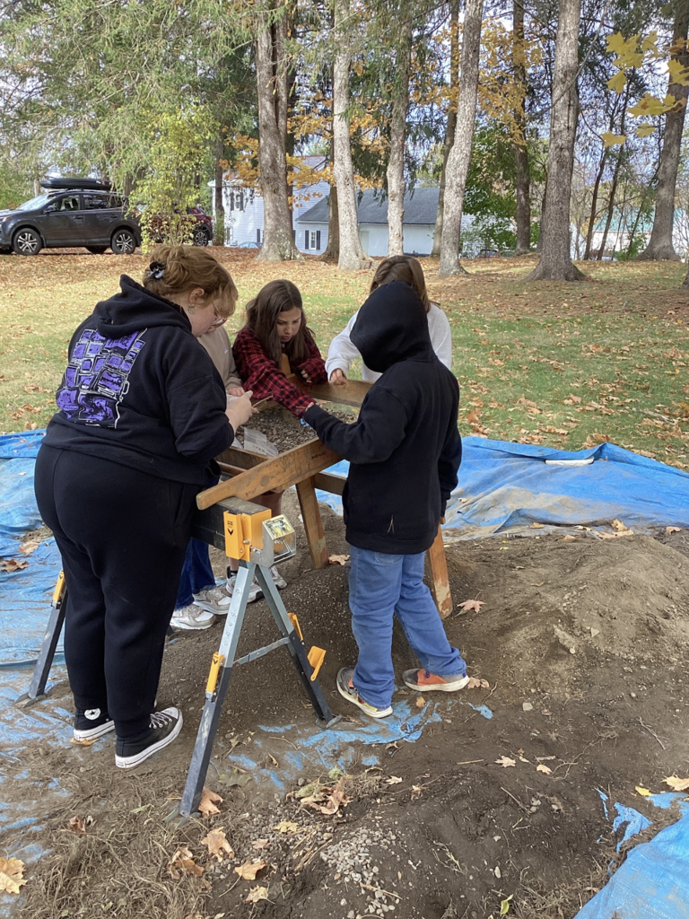 Students sift dirt through a wooden screen during an outdoor archaeology activity on a tarp-covered area.